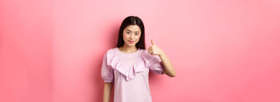 Smiling asian girl show thumb up in approval, praise good choice, like and recommend product, standing on pink background.