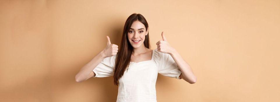 Very good. Happy and satisfied young woman showing thumbs-up, smiling positive, approve and like excellent choice, praising you, make well done gesture, beige background.