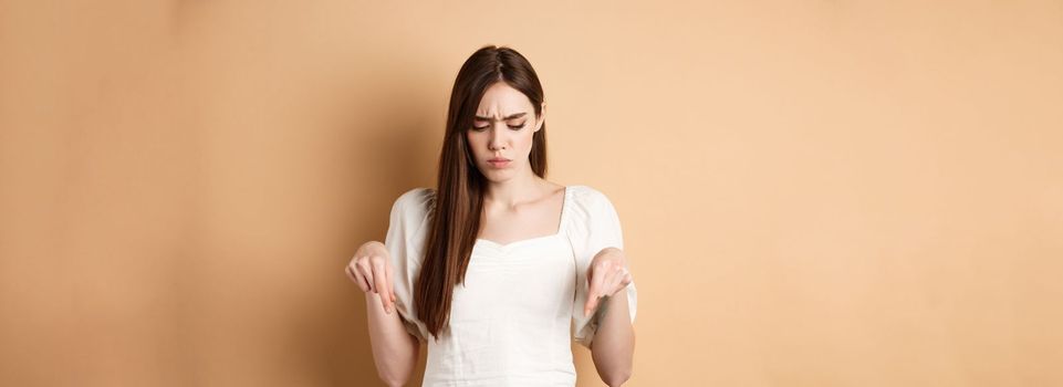 Upset young woman frowning and looking down, pointing at bad thing, standing confused and displeased on beige background.
