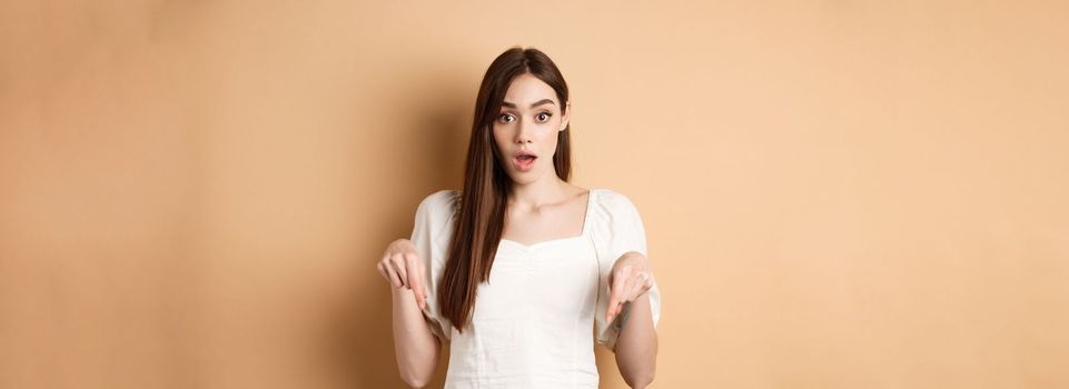 Wow look there. Amazed young woman in white dress pointing fingers down, standing in awe with dropped jaw and popped eyes against beige background.