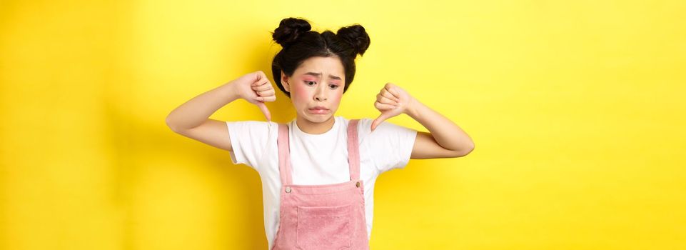 Asian beauty girl cringe from something disgusting, look away and showing thumbs-down to express dislike, negative emotions, standing in summer clothes on yellow background.