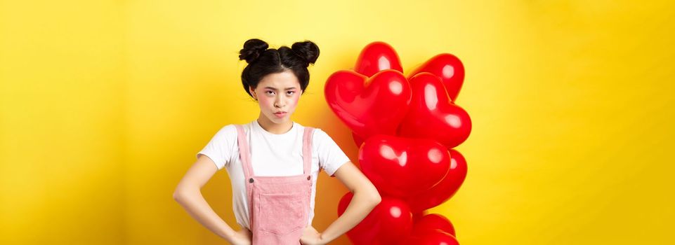 Valentines day and relationship concept. Disappointed asian girlfriend sulking, looking offended at camera, mad at boyfriend, standing near heart balloons, yellow background.