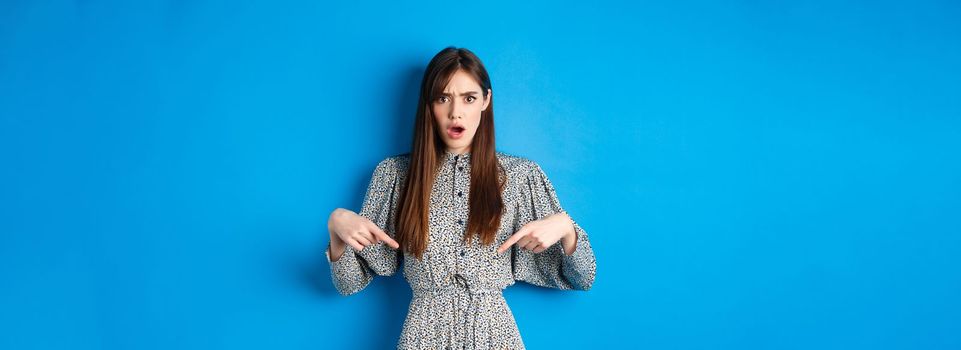 Shocked and offended young woman in dress frowning, gasping and pointing fingers down at insulting promo, standing on blue background.