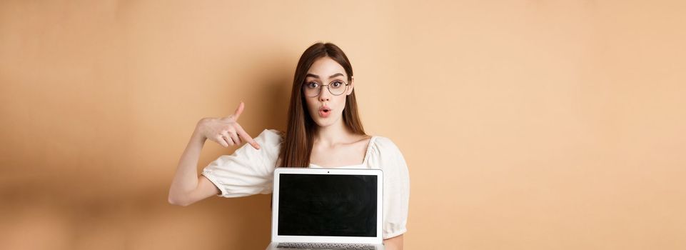 Excited girl in glasses making presentation on computer, pointing hand at laptop screen and say wow, standing on beige background.