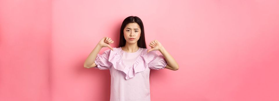 Skeptical and disappointed asian woman showing thumbs down with reluctant smirk, dislike product, show negative emotion, standing on pink background.