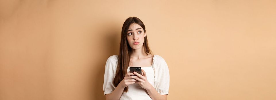 Confused young woman thinking after reading news on mobile phone, looking at upper left corner hesitant, standing on beige background.