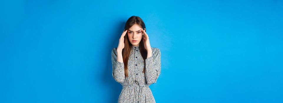 Woman trying to concentrate, touching head and squinting at camera, thinking or remember something, standing in dress on blue background.