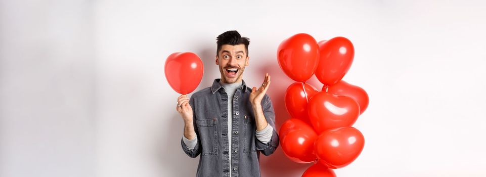 Valentines day concept. Cheerful young man celebrating love holiday, standing near red heart balloons and looking surprised, scream of joy, white background.