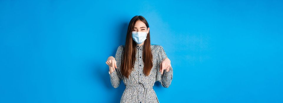 Covid-19, social distancing and healthcare concept. Happy beautiful person in medical mask pointing fingers down, laughing and smiling, showing advertisement, blue background.
