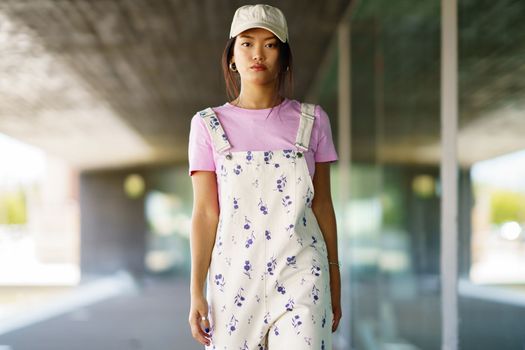 Young Asian female in stylish outfit and cap looking at camera and walking on blurred background of modern building on sunny day in city
