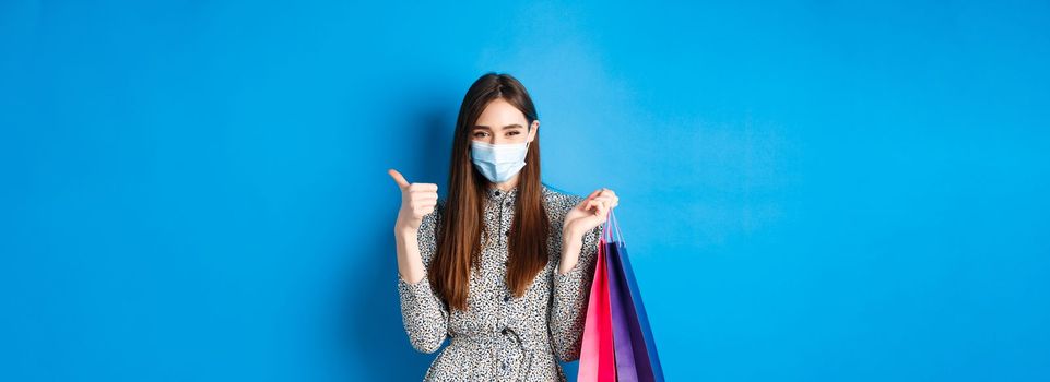 Covid-19, pandemic and lifestyle concept. Attractive woman wear medical mask on shopping, show thumb up and hold bags with purchases, blue background.