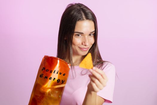 Positive young female model demonstrating package of nacho chips with Positive Energy inscription while looking at piece near pink background
