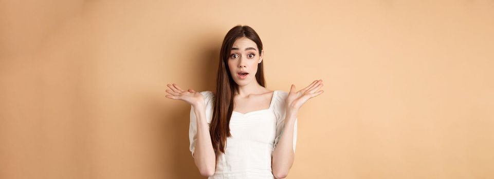 Surprised caucasian girl look with disbelief and amazement, spread hands sideways and gasping confused, cant understand what happening, know nothing, standing on beige background.