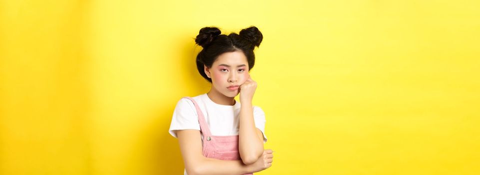 Bored asian teen girl with stylish makeup and summer clothes, looking reluctant and indifferent, standing on yellow background.