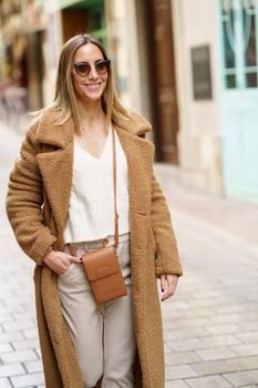 Confident female in sunglasses and brown coat with hand in pocket smiling while walking on town street