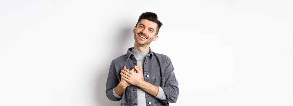 Romantic guy hold hands on heart and smiling, thanking you, looking pleased and grateful, standing on white background.