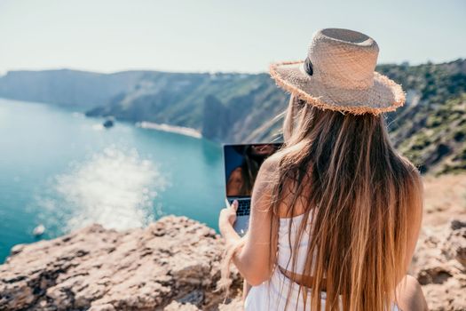 Successful business woman in yellow hat working on laptop by the sea. Pretty lady typing on computer at summer day outdoors. Freelance, travel and holidays concept.