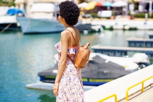 African American female tourist in summer dress with backpack and sunglasses standing on embankment and admiring sea water on sunny weekend day in harbor