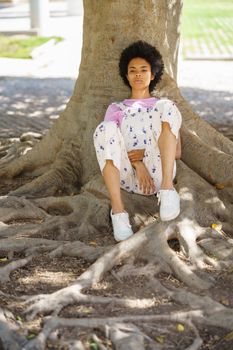 Full body of dreamy African American female looking away while sitting near thick tree trunk with roots in park on summer day