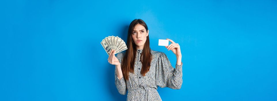 Shopping. Thoughtful girl looking up and biting lip, counting in mind or thinking, holding plastic credit card with dollar bills, standing on blue background.