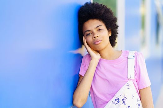 Calm young African American female in casual wear looking at camera and touching cheek while standing near blue building in city