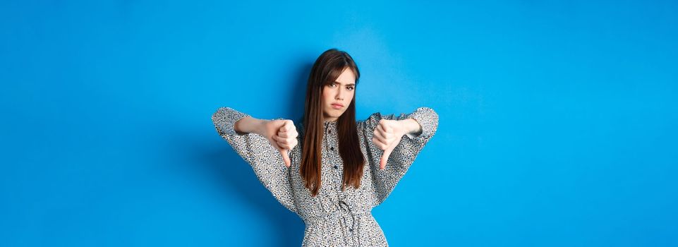 Negative emotion. Disappointed frowning woman with long hair and dress, showing thumbs down and grimacing with dislike, express aversion, standing on blue background.