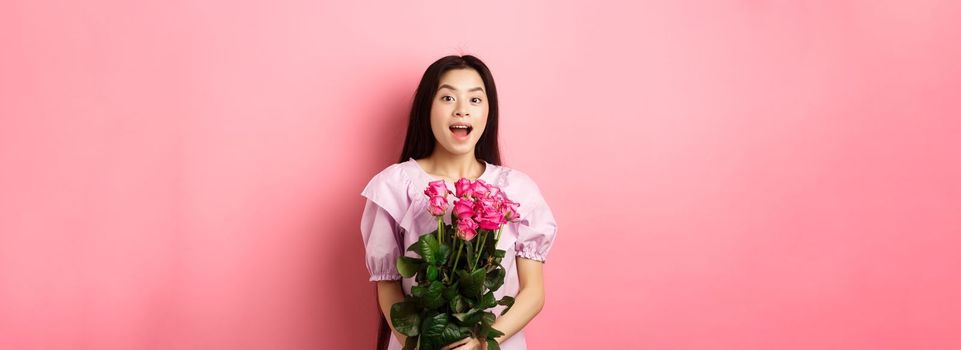 Korean teen girl in dress having romantic date on Valentines day, holding bouquet of roses and looking surprised at camera, receive gift on date from lover, pink background.