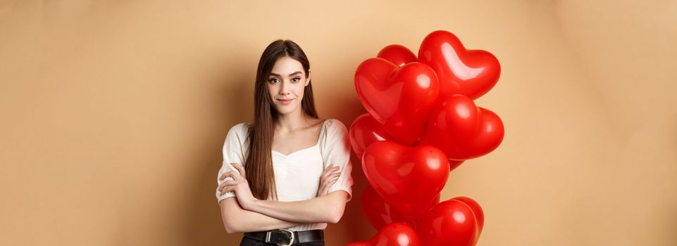 Sassy young girl cross arms on chest and smiling confident, standing near Valentines day hearts balloons and looking at camera, beige background.