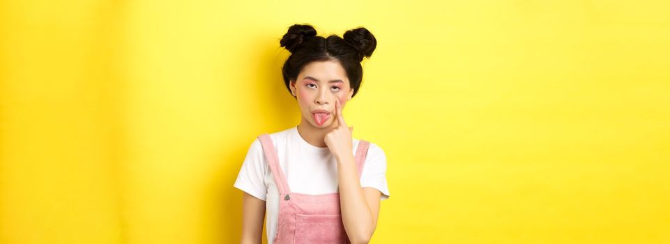 Rude asian girl stretch eyelid and showing tongue, mocking someone, standing on yellow background.