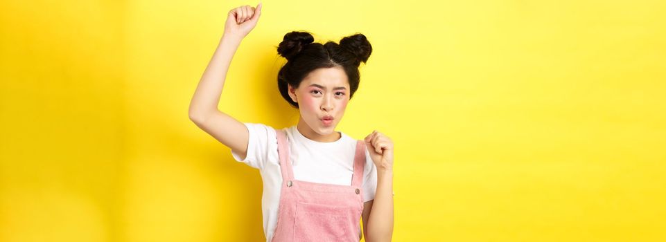 Excited asian woman getting motivation, raising hand up and chanting, celebrating victory, triumphing and standing on yellow background.