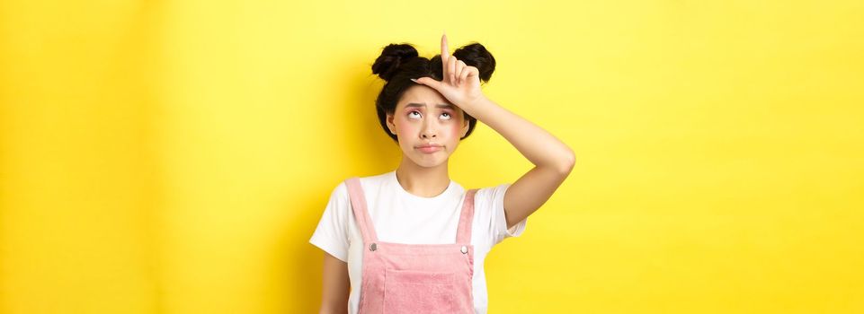 Sad girl showing loser sign on forehead and sulking upset, feeling disappointed in herself, standing on yellow background.