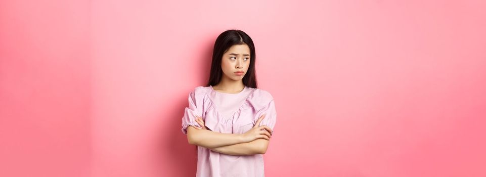 Sad and gloomy asian woman look away at logo, sulking from unfair situation, cross arms on chest disappointed, standing against pink background.