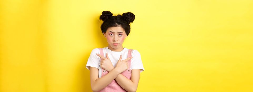 Indecisive timid asian girl pointing sideways, showing left and right ways and sulking upset, standing sad on yellow background.