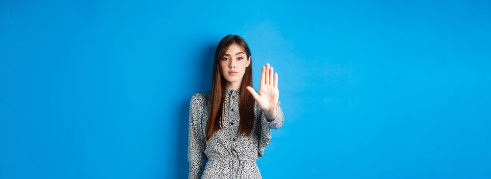 Serious and confident woman with long hair and dress, stretch out hand and say no, make stop gesture, prohibit bad action, standing on blue background.