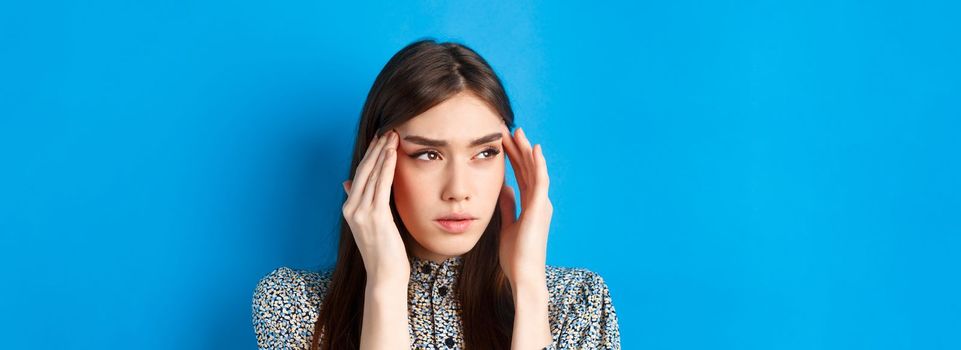 Close-up portrait of young woman feel sick, touching head temples and frowning from headache, having migraine, looking aside at logo, blue background.