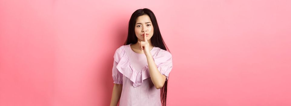 Cute asian girl tell to be quiet, scolding loud person, showing shush sign with finger pressed to lips, standing in dress on pink background.