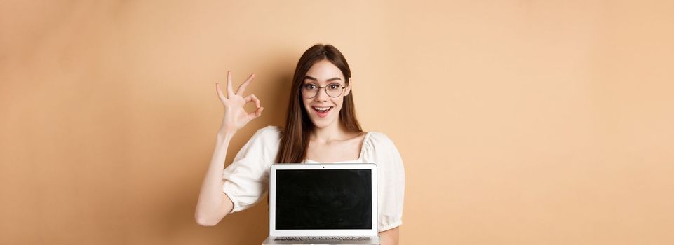 E-commerce. Excited young woman in glasses showing okay sign and laptop screen, recommending internet promo, standing on beige background.