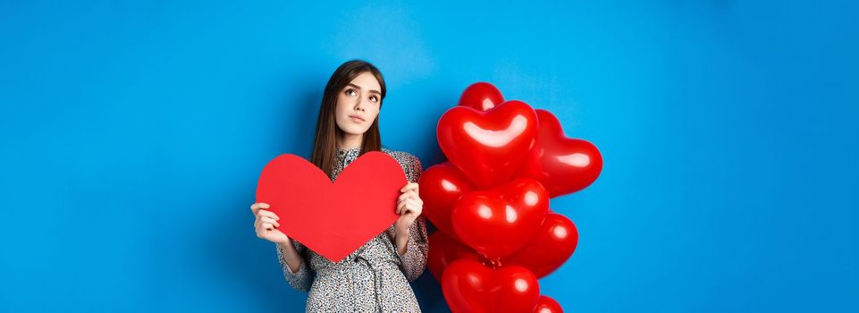 Valentines day. Dreamy pretty lady in dress, holding big red heart cutout and searching for true love, looking up pensive, standing near holiday balloons on blue background.