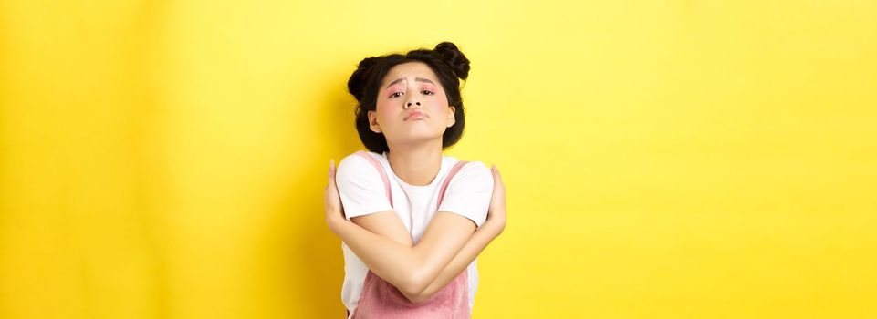 Lonely sad teen girl hugging herself, wanting to relationship and cuddles, standing alone on yellow background.