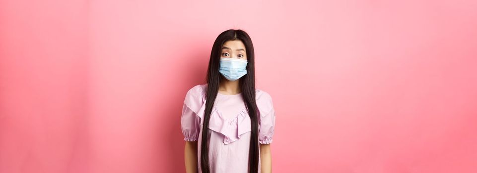 Covid-19, pandemic lifestyle concept. Surprised asian woman in medical mask look in awe, raising eyebrows with disbelief, standing against pink background.