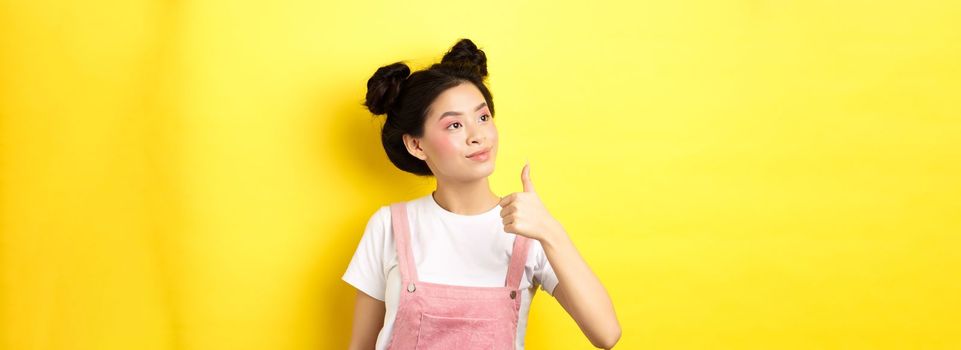 Glamour asian girl with pink makeup and summer clothes, looking left at logo with thumb up, approve and praise, standing on yellow background.