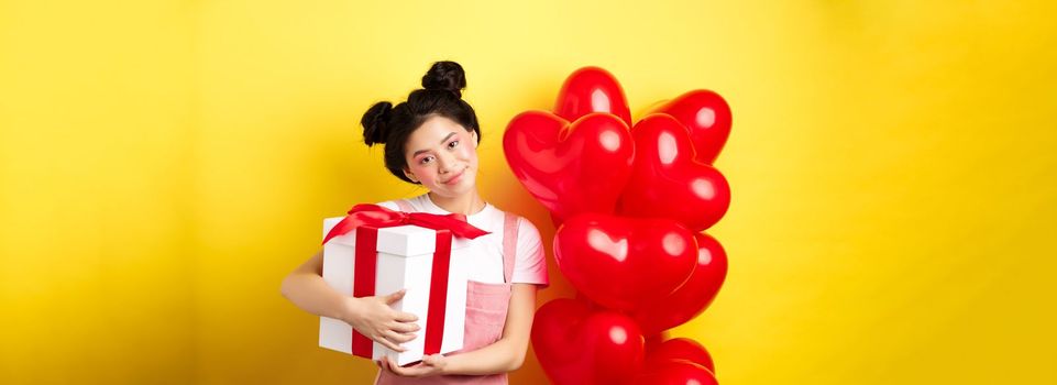 Happy Valentines day. Cute asian girl hugging surprise gift from boyfriend, standing near lovely red heart balloons and yellow background.