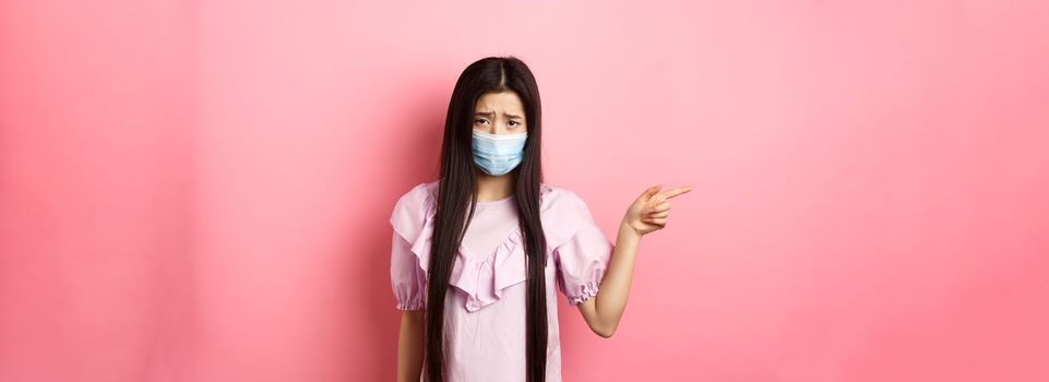 Covid-19, pandemic lifestyle concept. Sad and gloomy asian girl in medical mask pointing left, complain on unfair situation, standing disappointed against pink background.