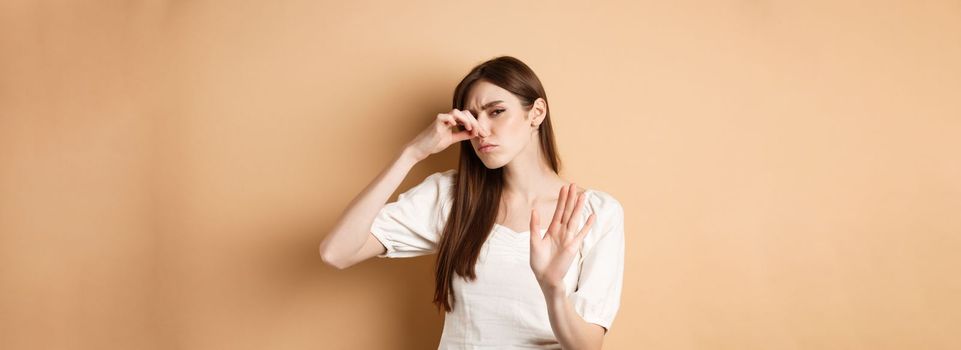 Displeased woman plug her nose from bad smell, showing stop gesture and frowning from disgust. Shut nostrils in aversion, standing on beige background.