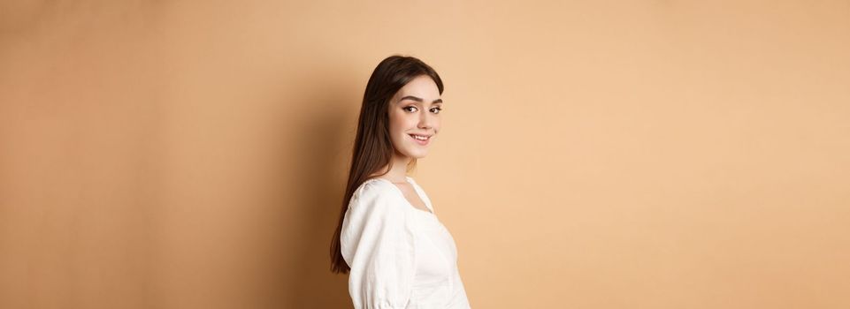 Profile of pretty european girl turn head at camera and smiling coquettish, standing in white blouse against beige background.