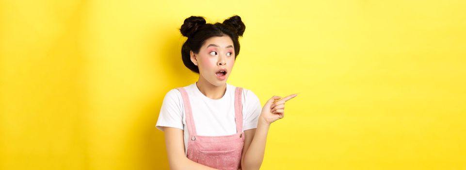 Intrigued asian beauty girl with makeup, looking curious at logo, pointing finger aside, standing in summer clothes against yellow background.