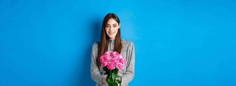 Valentines day concept. Tender young woman in dress, holding bouquet of roses on romantic date, standing on blue background.