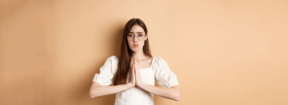 Sad girl in glasses begging for help, say please and looking cute, need favour, standing on beige background.