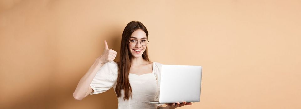 Smiling woman in glasses using laptop and showing thumb up, working on computer, standing on beige background.