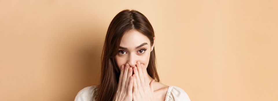 Close up of coquettish pretty girl covering mouth, looking happy and romantic, standing on beige background.